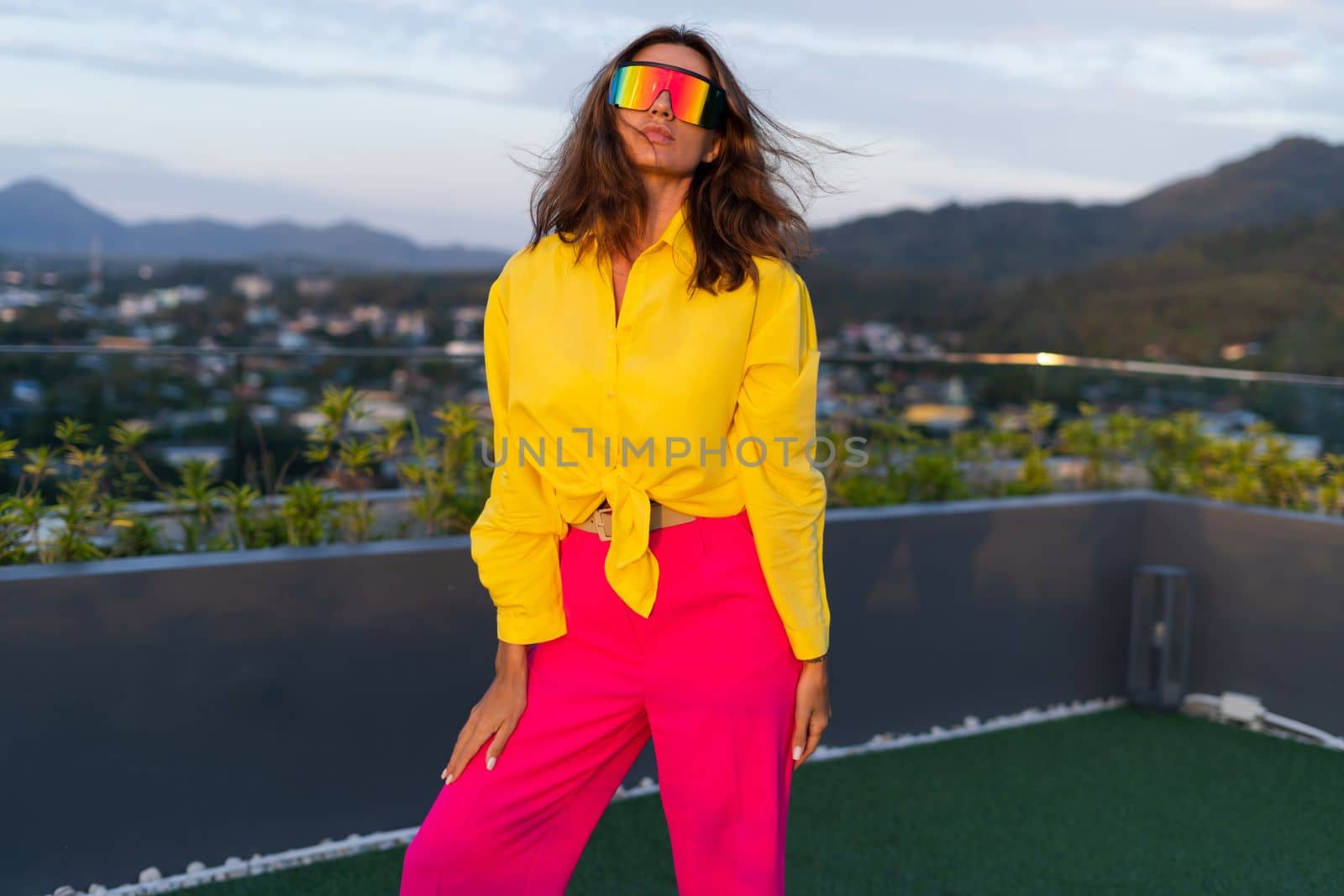 Stylish fit fashion women in bright pink wide leg pants and yellow shirt holding bag trendy sunglasses posing at rooftop terrace tropical view outdoor natural day light by kroshka_nastya