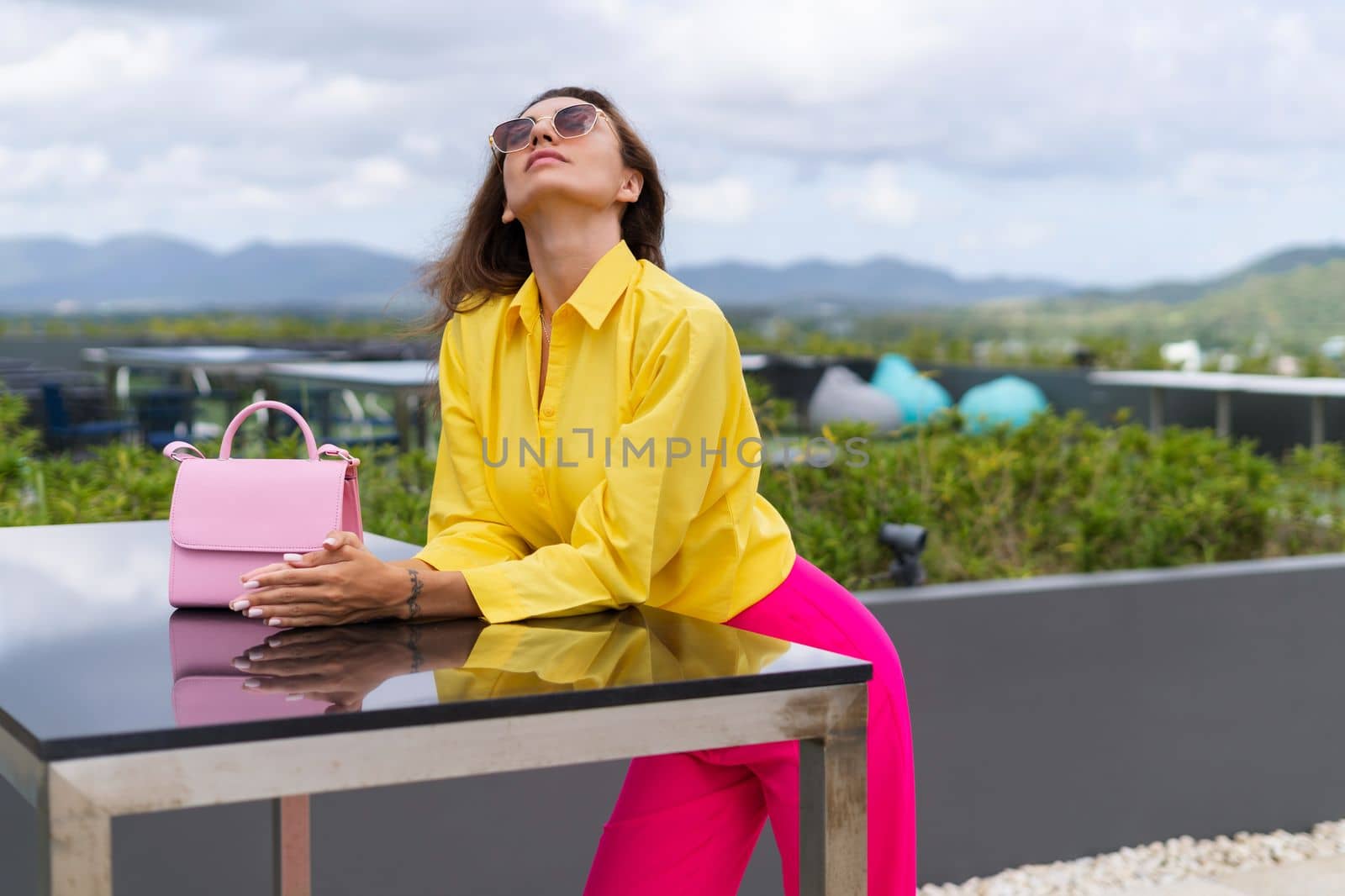 Stylish fit fashion women in bright pink wide leg pants and yellow shirt holding bag trendy sunglasses posing at rooftop terrace tropical view outdoor natural day light by kroshka_nastya