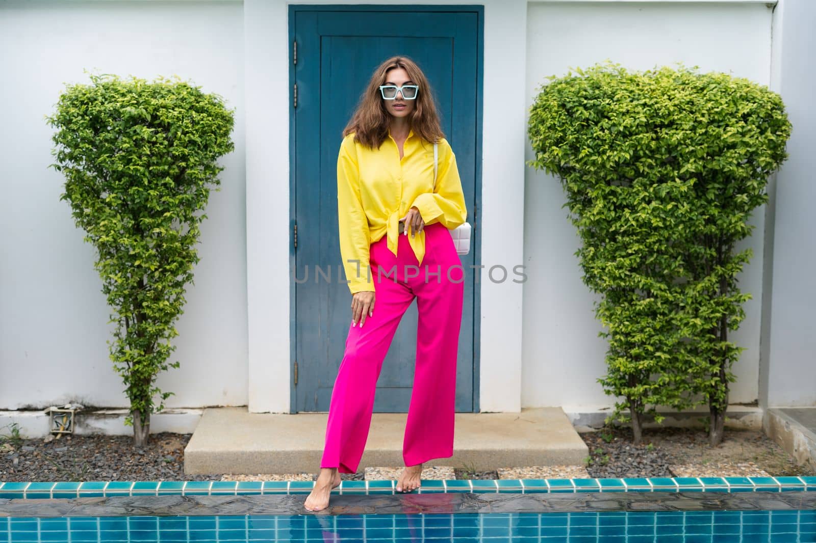 Stylish fit fashion women in bright pink wide leg pants and yellow shirt holding bag trendy mint sunglasses posing at luxury tropical villa by pool outdoor natural day light by kroshka_nastya