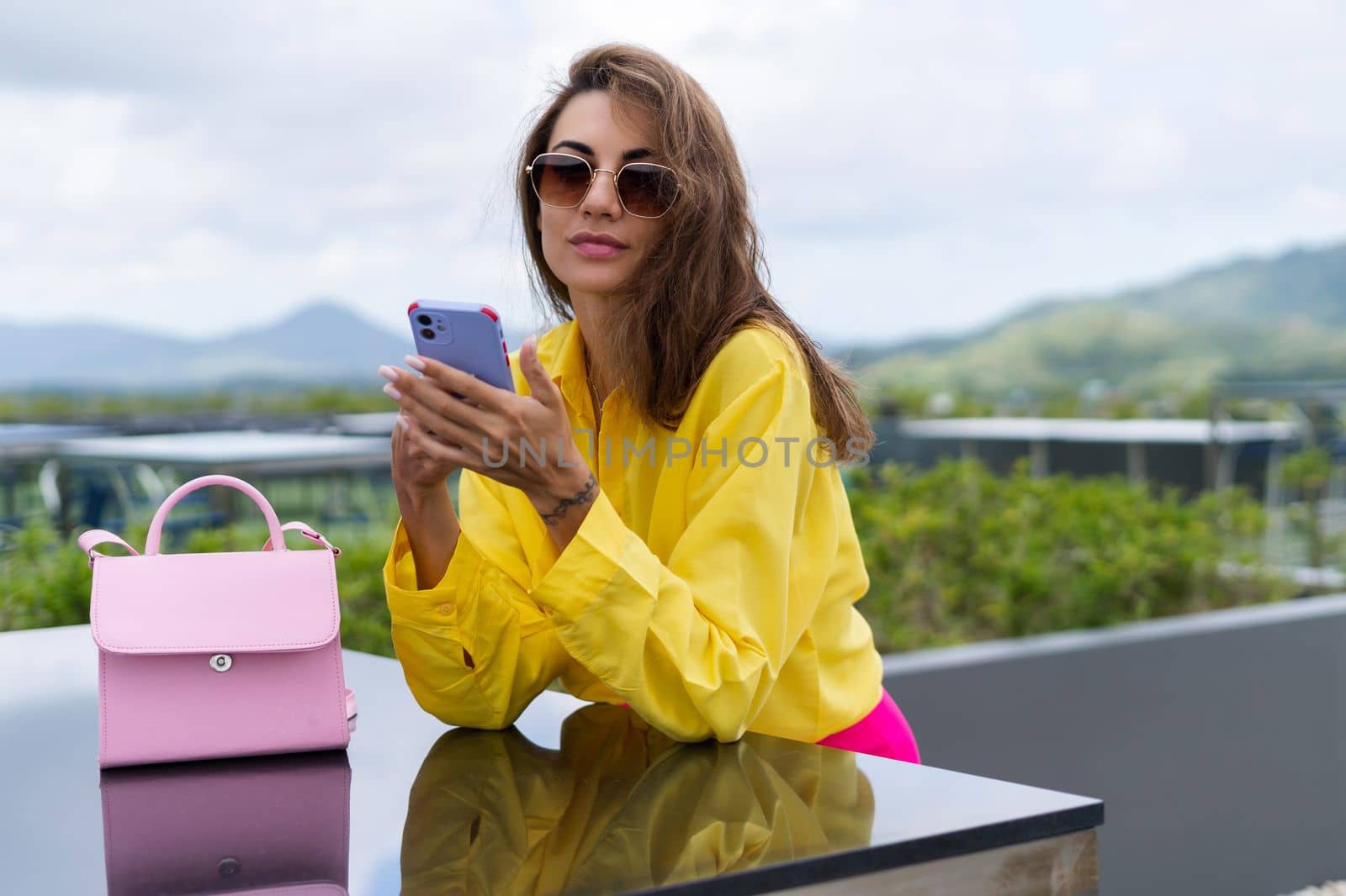 Stylish fit fashion women in bright yellow shirt trendy sunglasses posing at rooftop terrace tropical view outdoor holding mobile phone by kroshka_nastya