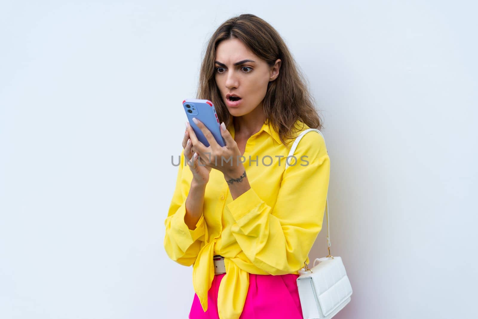 Stylish fit fashion women in yellow shirt and bright pink pants trendy posing on white wall background outdoor natural day light looking at phone screen confused shocked by kroshka_nastya