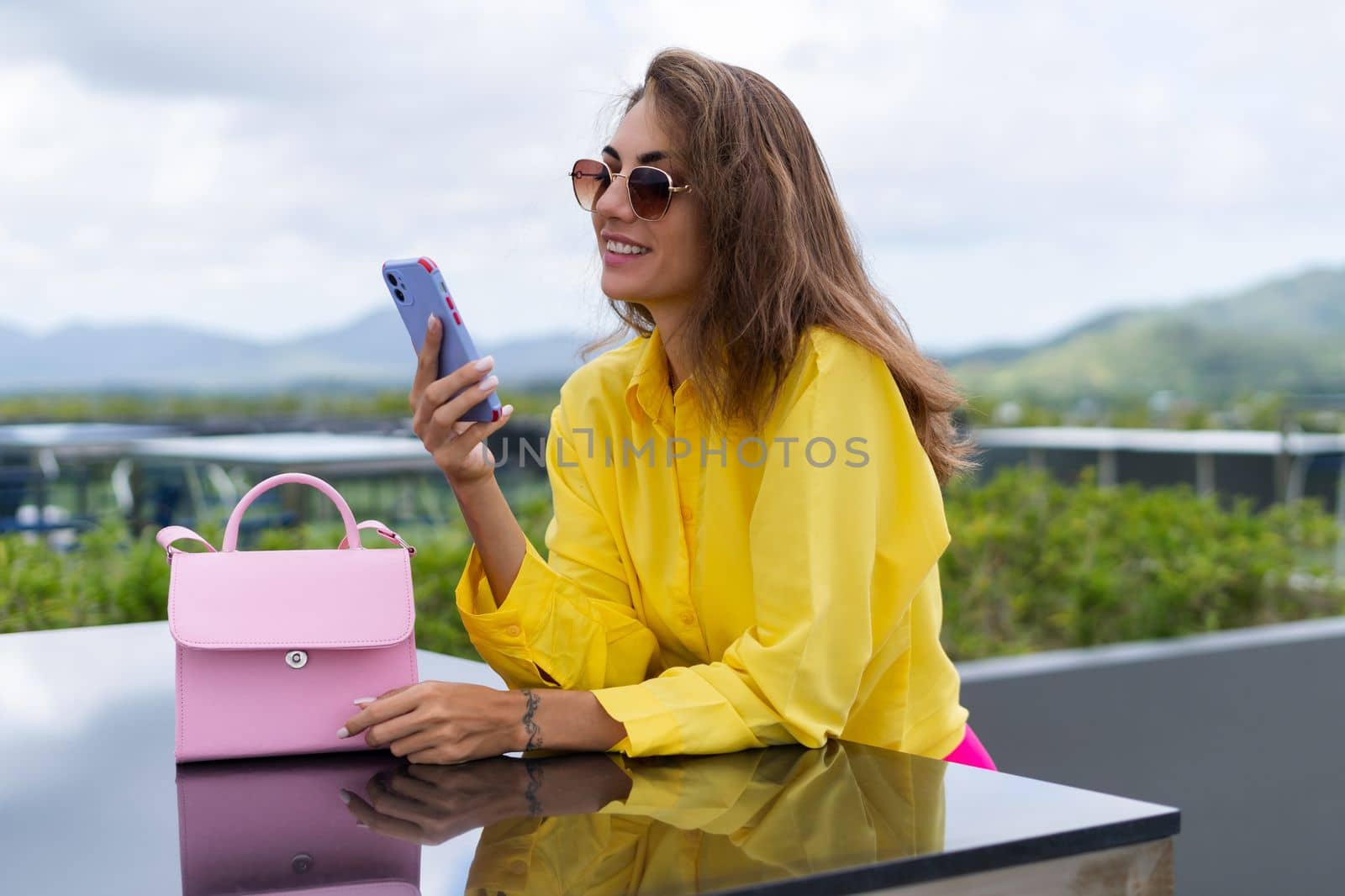 Stylish fit fashion women in bright yellow shirt trendy sunglasses posing at rooftop terrace tropical view outdoor holding mobile phone by kroshka_nastya