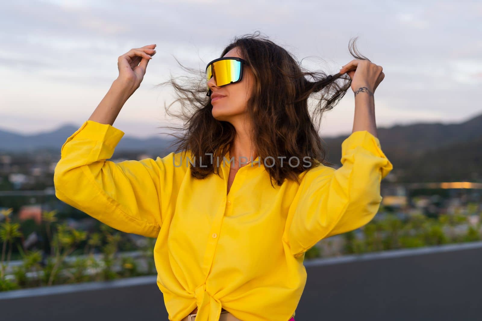 Stylish fit fashion women in bright yellow shirt trendy shield visor rainbow sunglasses posing at rooftop terrace tropical view outdoor sunset warm light by kroshka_nastya