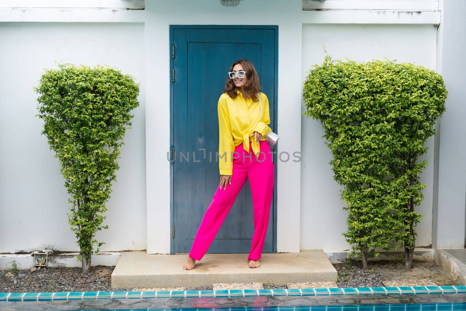 Stylish fit fashion women in bright pink wide leg pants and yellow shirt holding bag trendy mint sunglasses posing at luxury tropical villa by pool outdoor natural day light