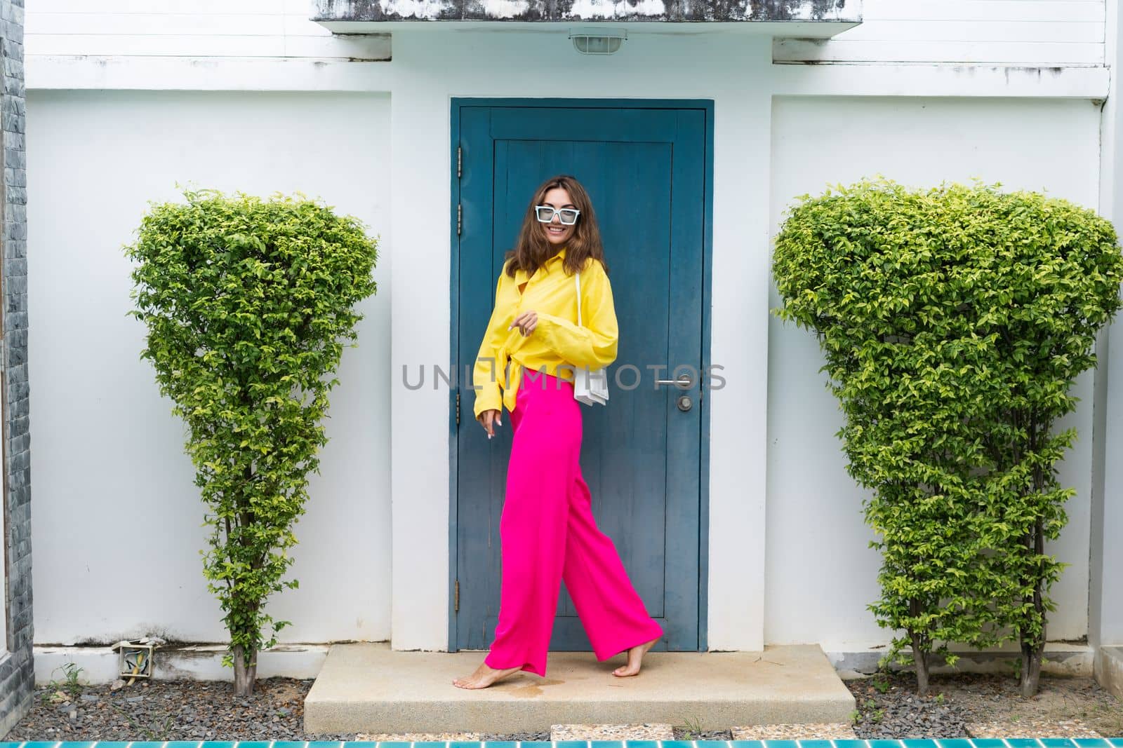 Stylish fit fashion women in bright pink wide leg pants and yellow shirt holding bag trendy mint sunglasses posing at luxury tropical villa by pool outdoor natural day light