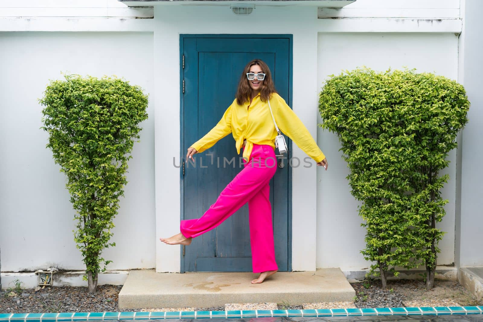 Stylish fit fashion women in bright pink wide leg pants and yellow shirt holding bag trendy mint sunglasses posing at luxury tropical villa by pool outdoor natural day light