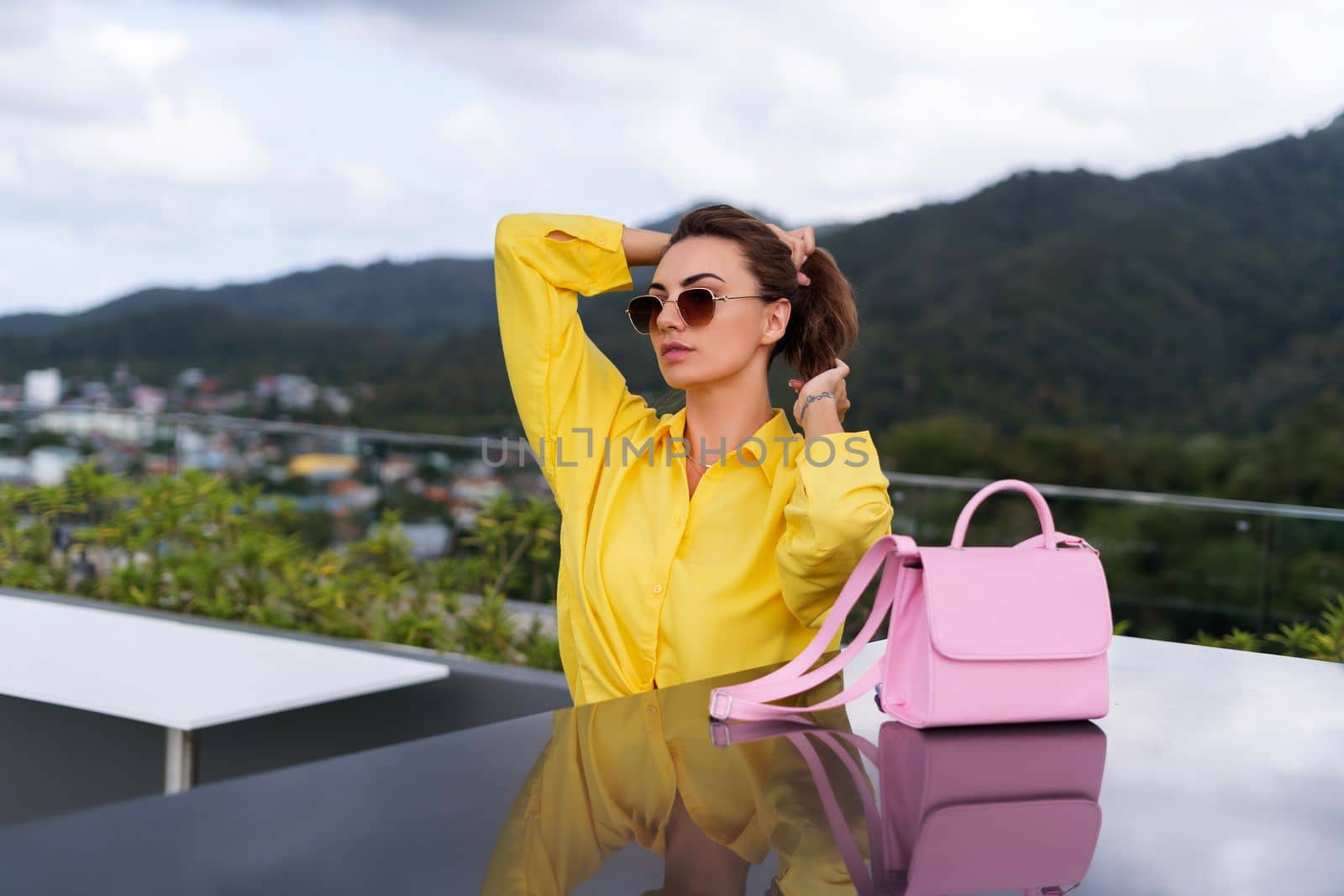 Stylish fit fashion women in bright yellow shirt trendy sunglasses posing at rooftop terrace tropical view outdoor with pink bag by kroshka_nastya