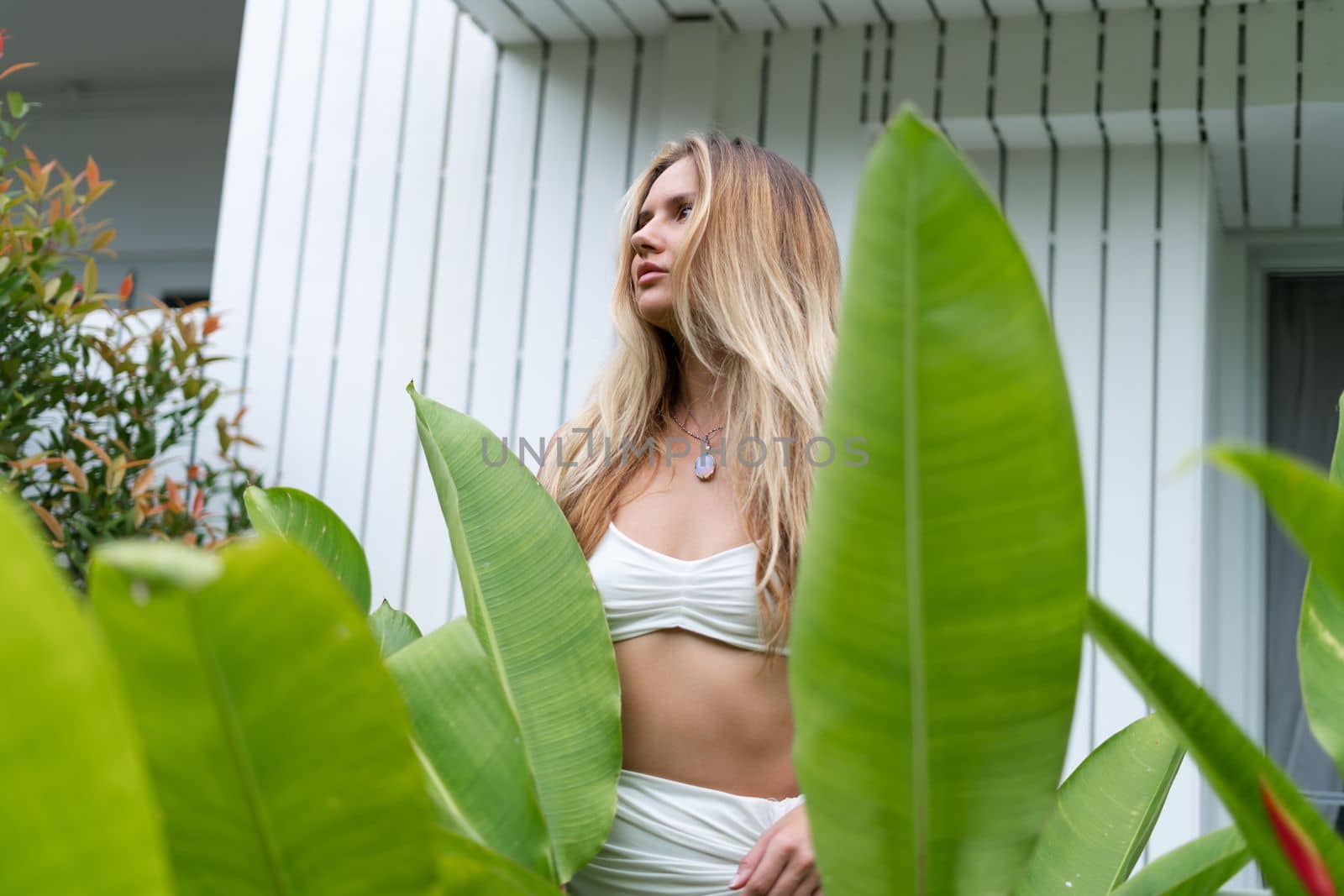 Beautiful young blonde woman in a top with long hair, natural beauty, daylight, in blue glasses, posing against the backdrop of a wooden fence and palm leaves