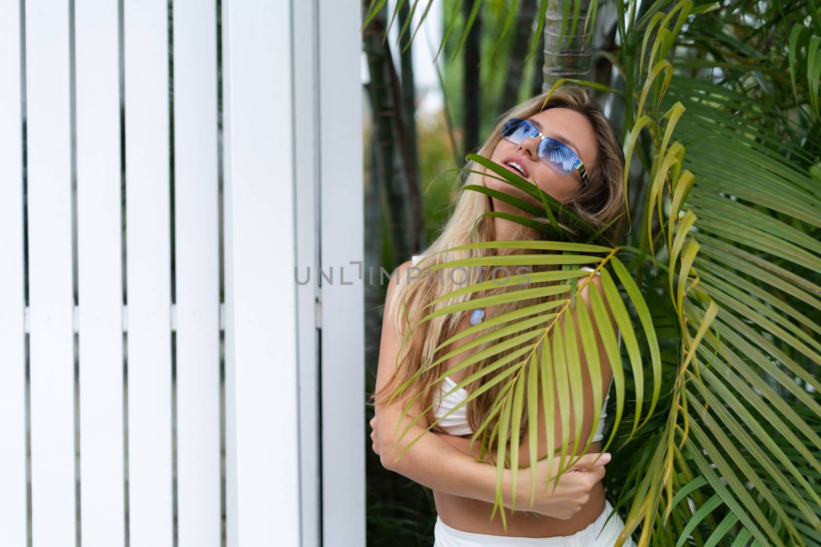 Beautiful young blonde woman in a top with long hair, natural beauty, daylight, in blue glasses, posing against the backdrop of a wooden fence and palm leaves