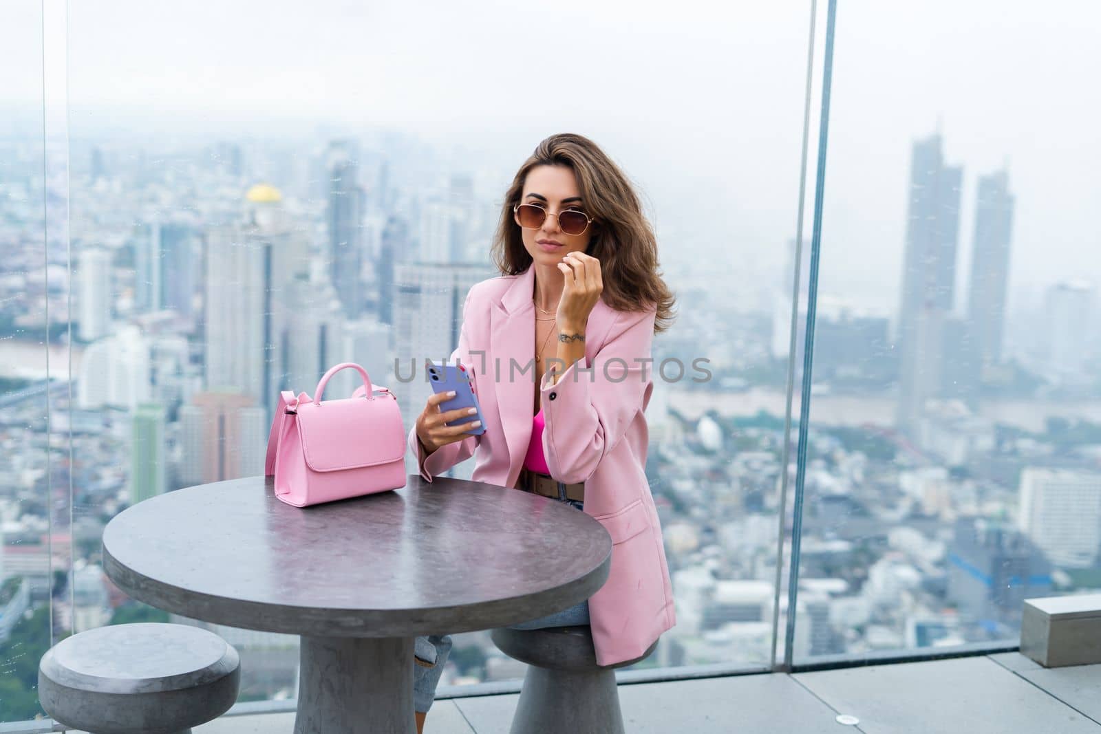 Stylish woman in beautiful fashion pink blazer with cute bag holding mobile phone on rooftop terrace with amazing Bangkok view high floor industrial city