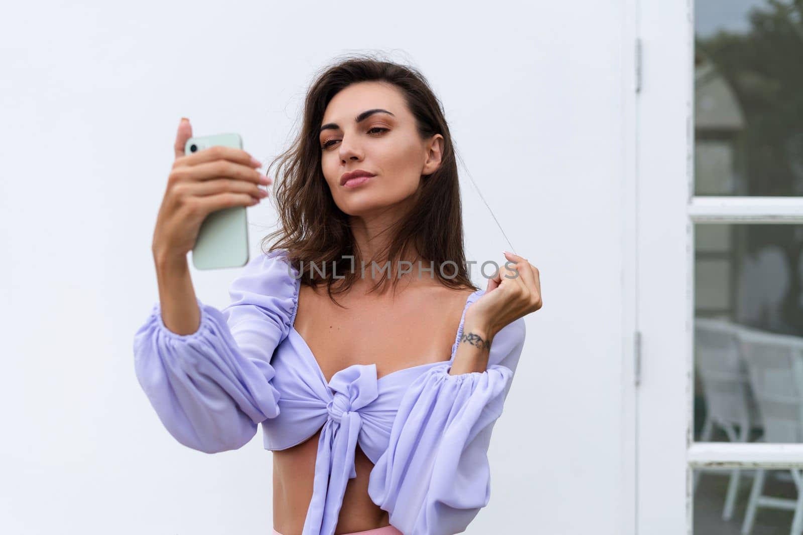 Pretty young stylish woman in trendy purple crop top long sleeve, isolated, white wall, natural day light..Taking selfie,look at mobile phone screen with smile. by kroshka_nastya