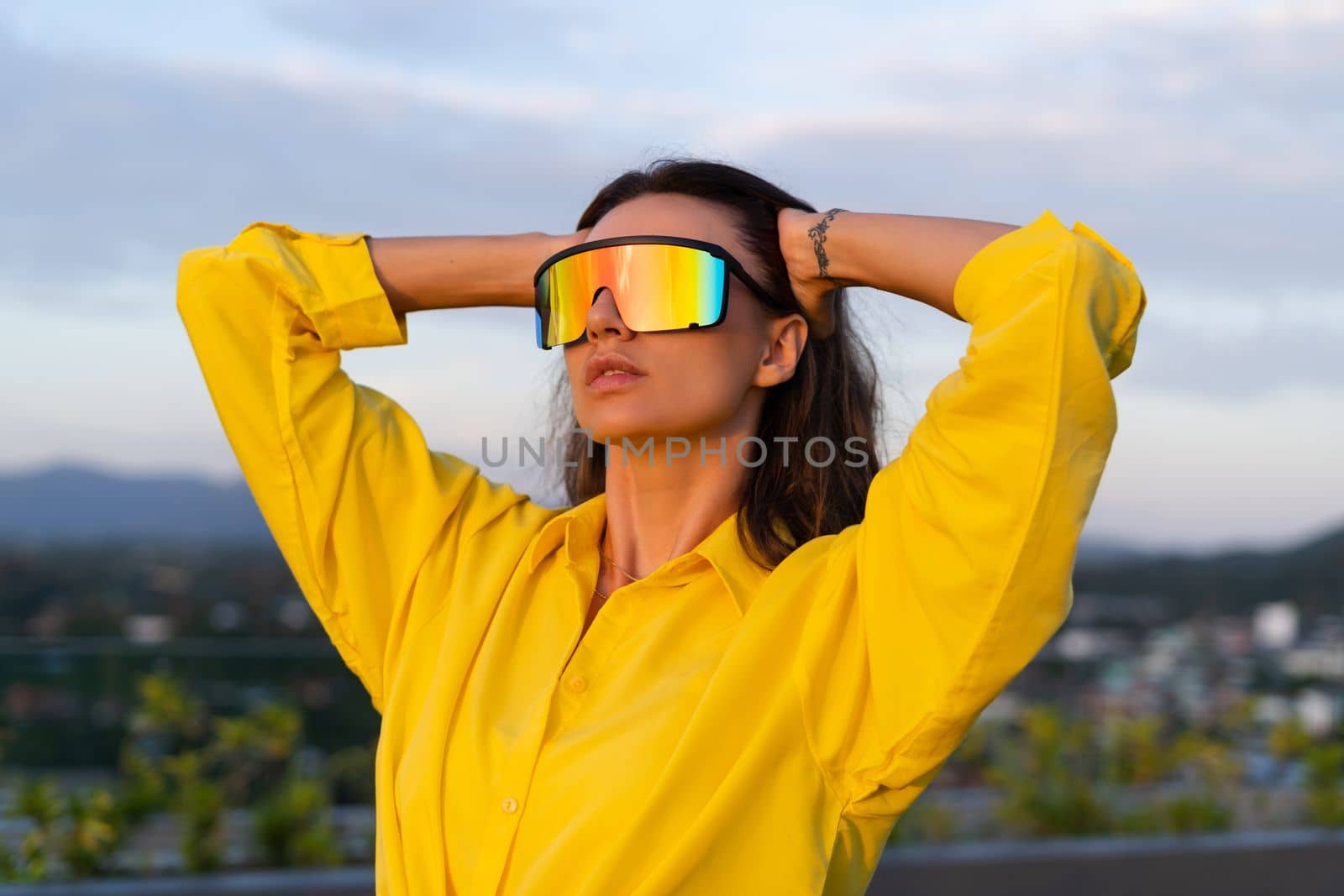 Stylish fit fashion women in bright yellow shirt trendy shield visor rainbow sunglasses posing at rooftop terrace tropical view outdoor sunset warm light by kroshka_nastya