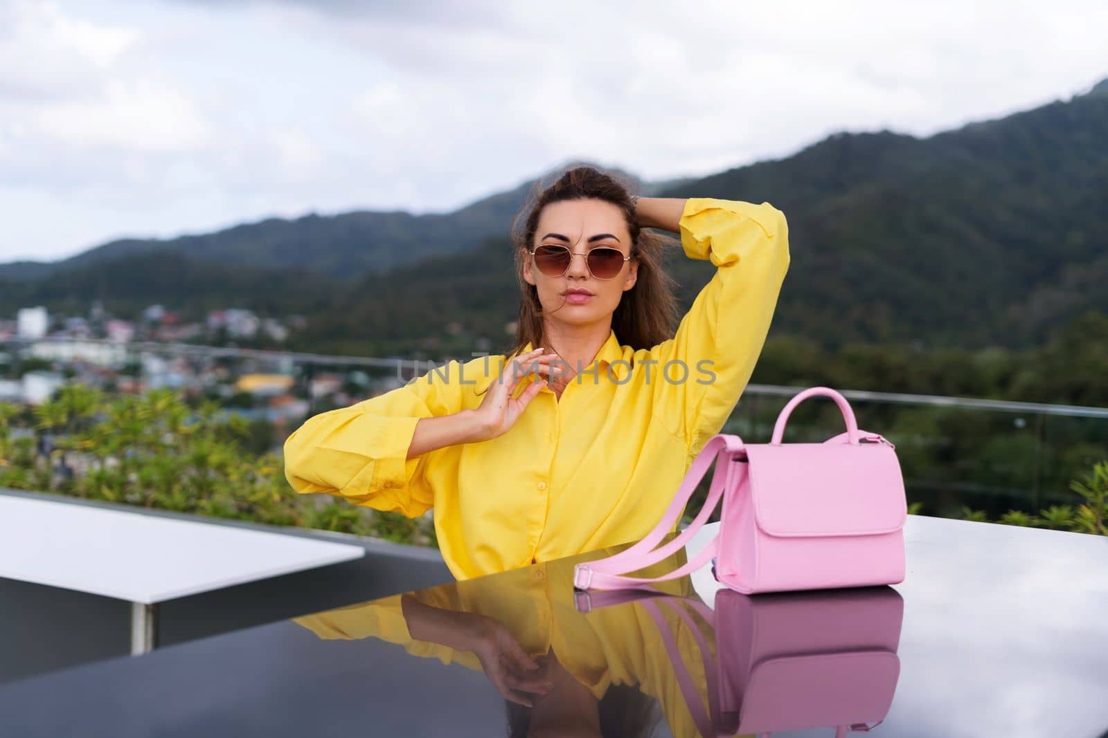 Stylish fit fashion women in bright yellow shirt trendy sunglasses posing at rooftop terrace tropical view outdoor with pink bag