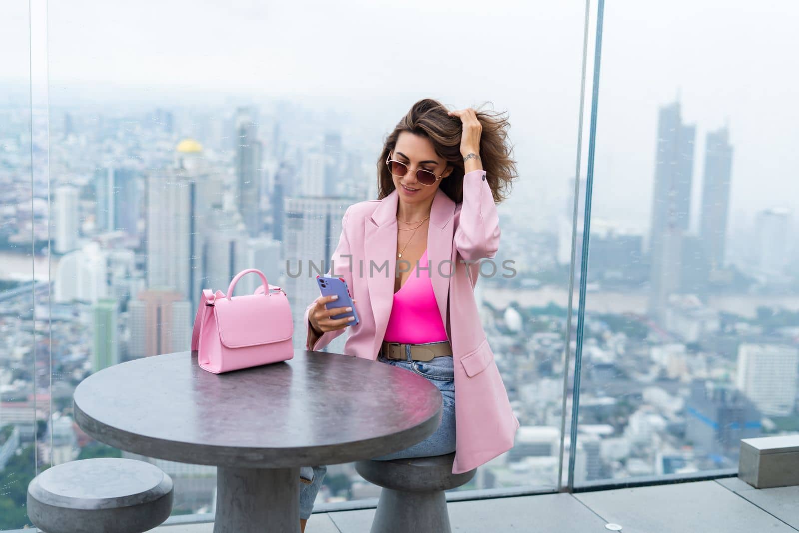 Stylish woman in beautiful fashion pink blazer with cute bag holding mobile phone on rooftop terrace with amazing Bangkok view high floor industrial city by kroshka_nastya