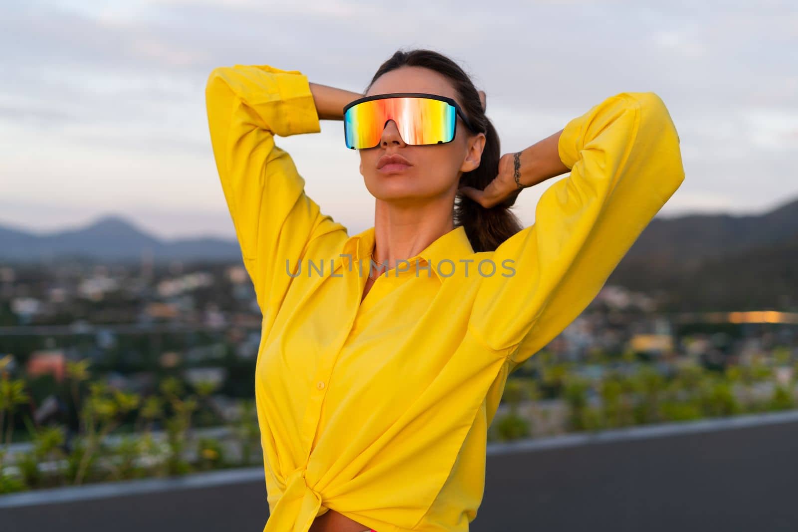 Stylish fit fashion women in bright yellow shirt trendy shield visor rainbow sunglasses posing at rooftop terrace tropical view outdoor sunset warm light