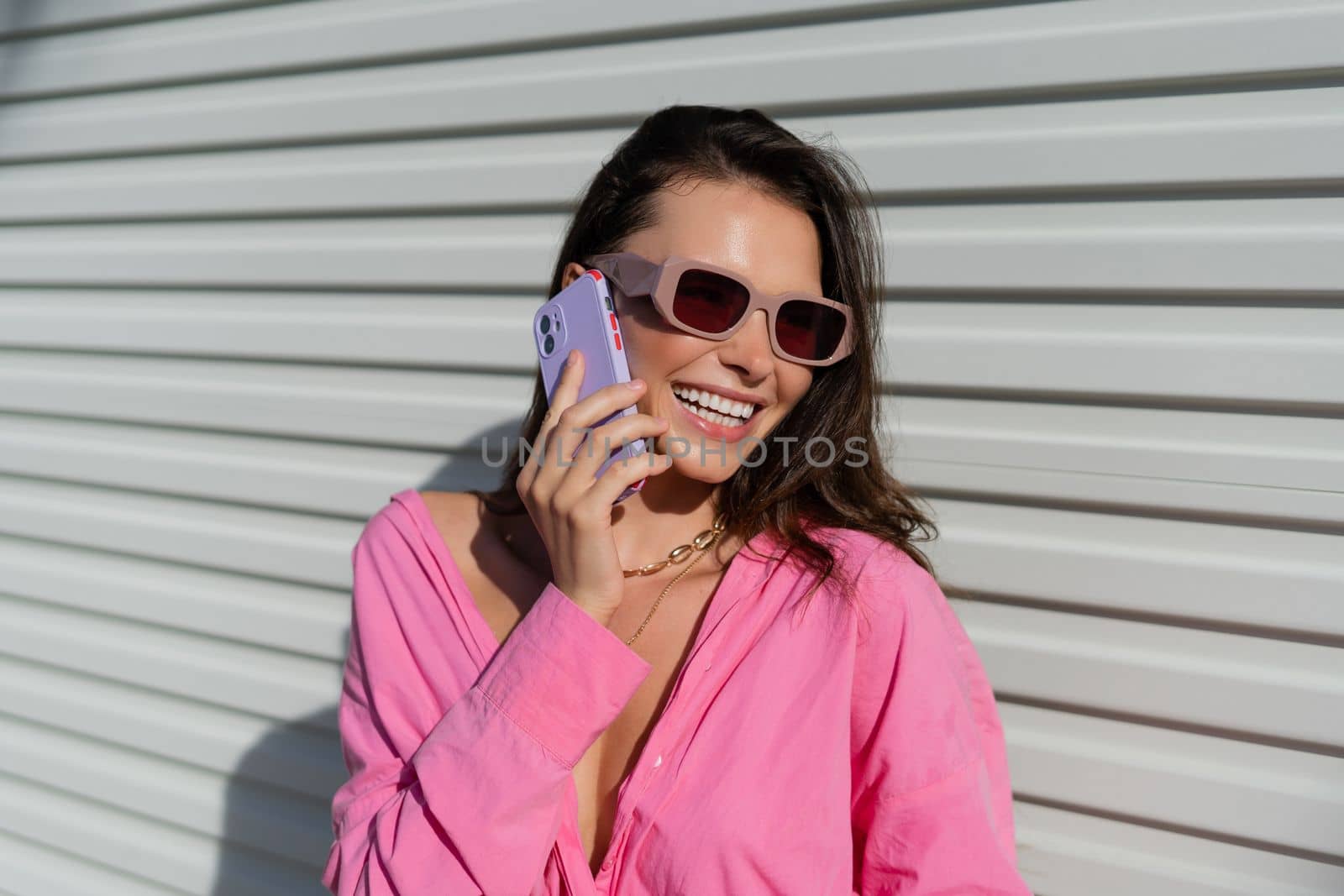 Young beautiful brunette woman in a pink shirt, neck jewelry, necklace, trendy sunglasses on the background of a light garage fence, calls the phone, laughs, smiles