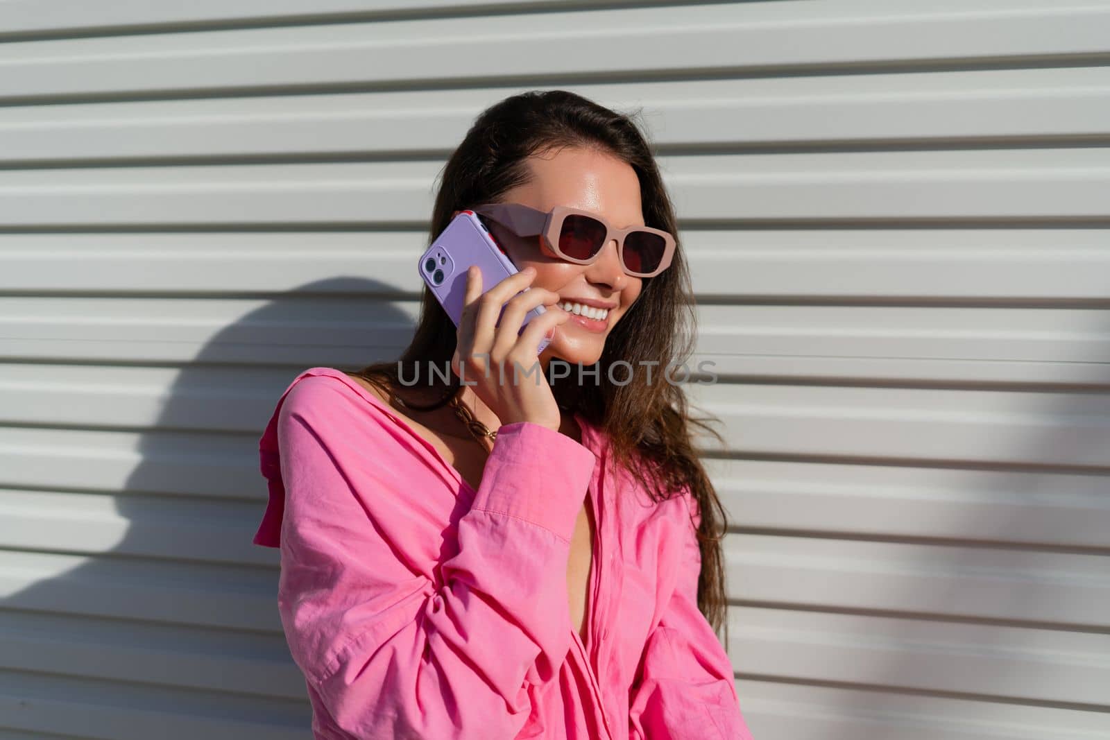 Young beautiful brunette woman in a pink shirt, neck jewelry, necklace, trendy sunglasses on the background of a light garage fence, calls the phone, laughs, smiles by kroshka_nastya