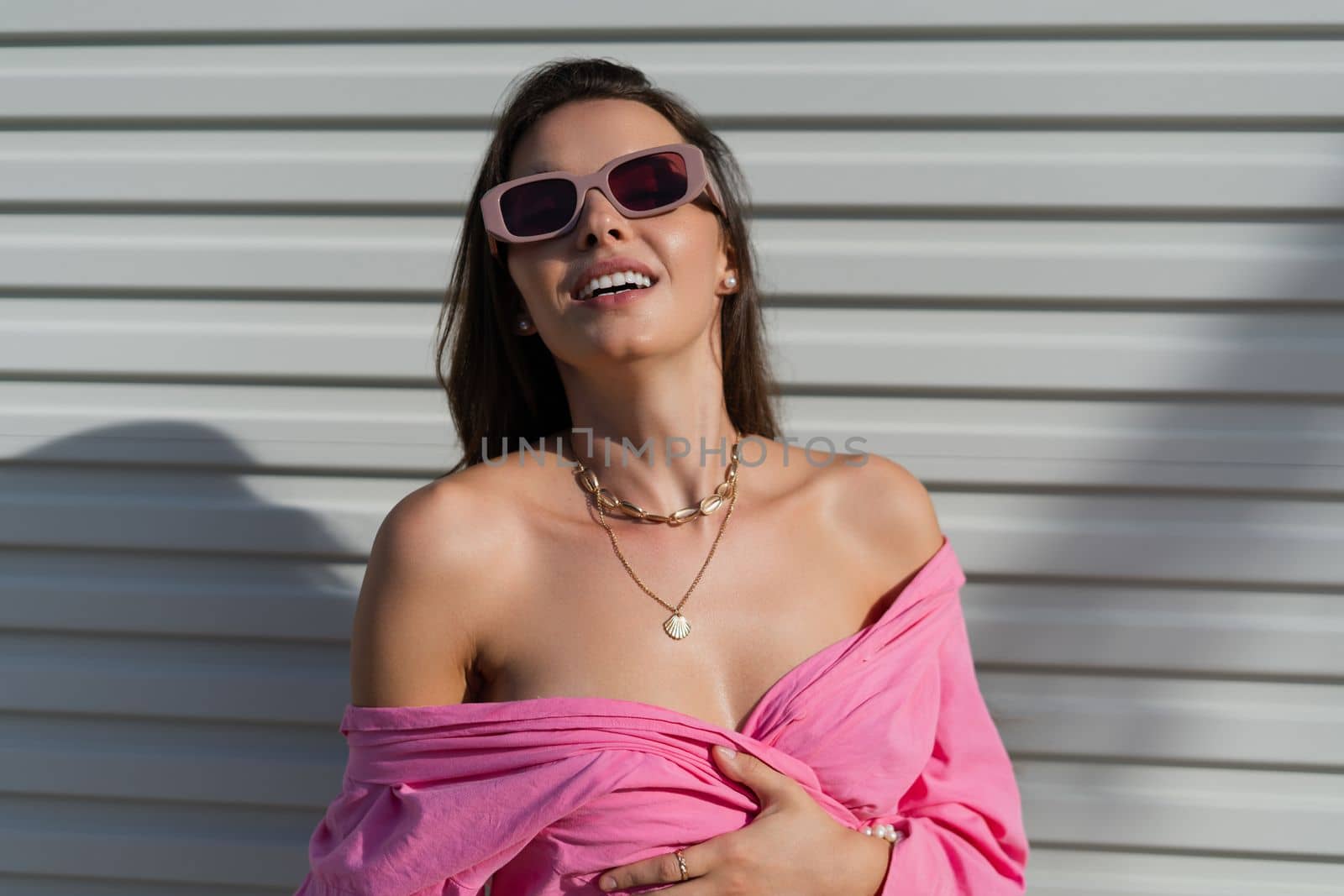 Young beautiful brunette in a pink shirt, neck jewelry, necklace, trendy sunglasses on the background of a light garage door fence