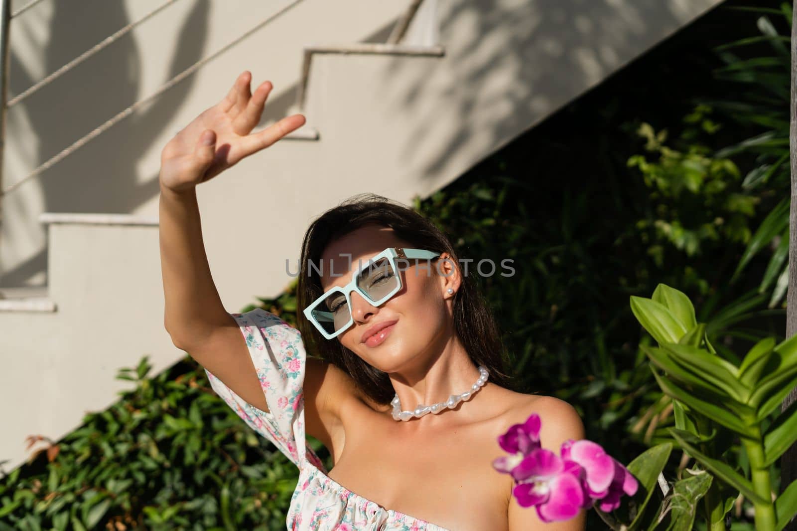 Young beautiful woman in a romantic dress with a floral print, sunglasses and a pearl necklace bracelet, against the backdrop of tropical leaves by kroshka_nastya