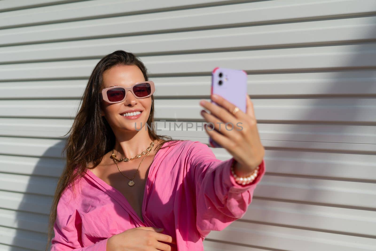 Young beautiful brunette in a pink shirt, neck jewelry, necklace, trendy sunglasses on the background of a light garage fence, takes a selfie on the phone, laughs, smiles by kroshka_nastya