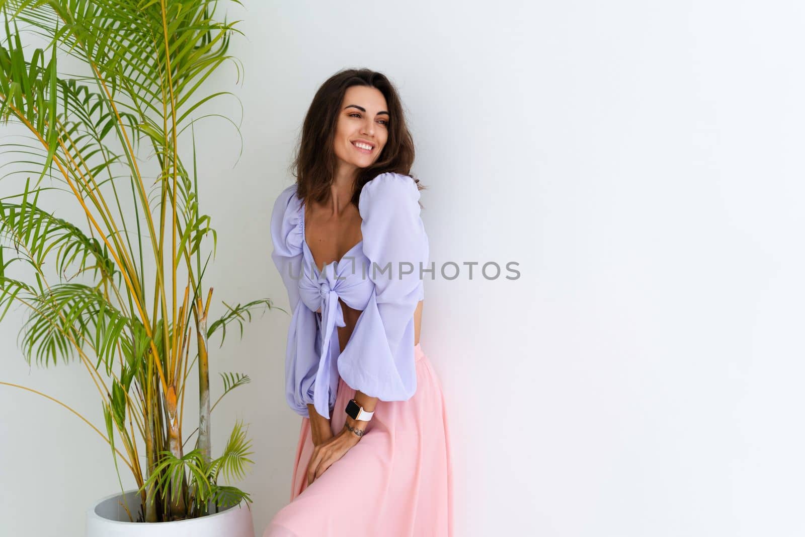 Stylish young woman with voluminous hair in a trendy long sleeve crop top and a pink skirt posing against a white wall and a tropical bush, wearing a smart watch by kroshka_nastya