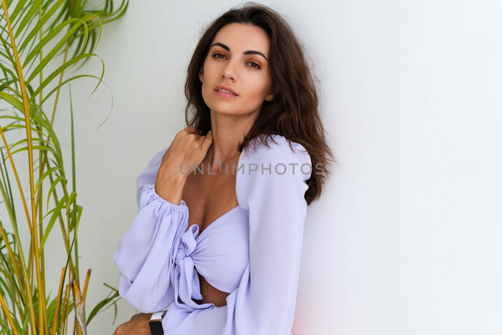 Stylish young woman with voluminous hair in a trendy long sleeve crop top and a pink skirt posing against a white wall and a tropical bush, wearing a smart watch by kroshka_nastya