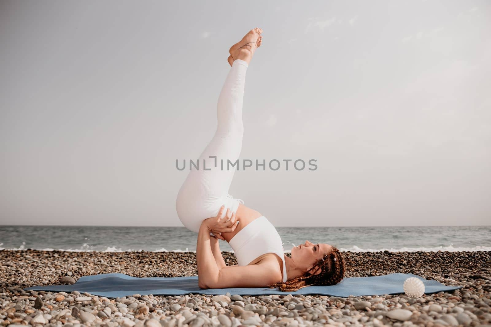 Woman yoga sea. Well looking middle aged woman with braids dreadlocks in white leggings and tops doing stretching pilates on yoga mat near sea. Female fitness yoga routine concept. Healthy lifestyle