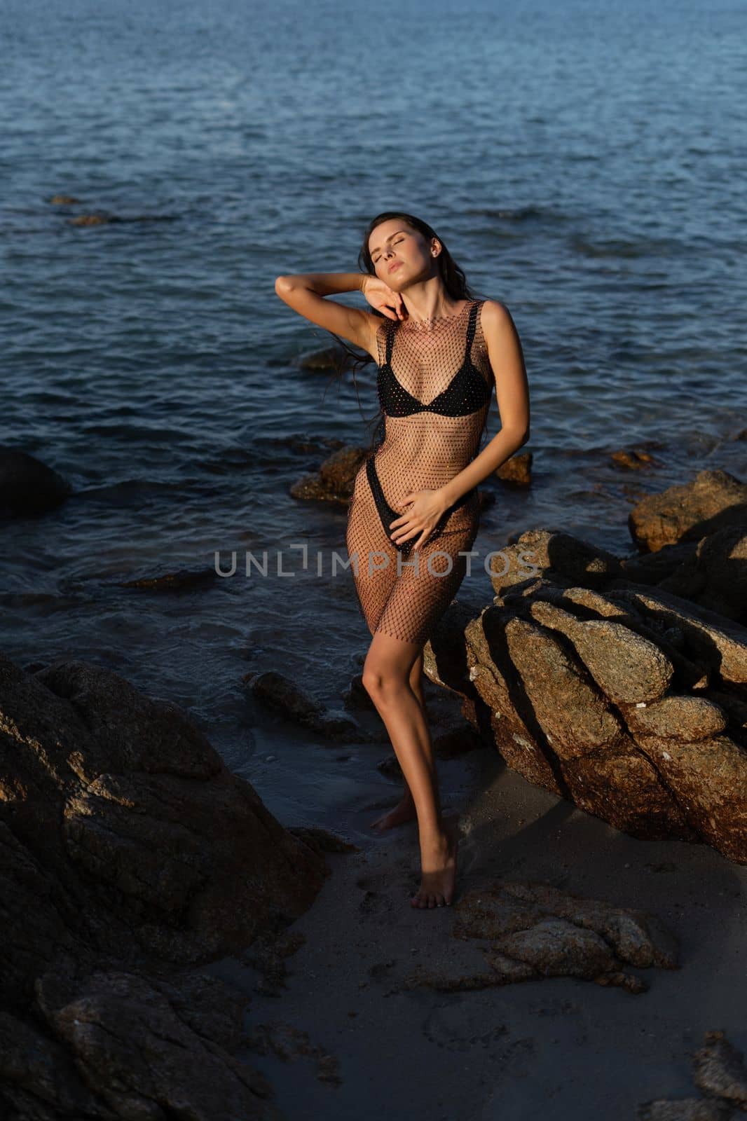 Young beautiful tanned woman on the background of the sea at sunset in a black swimsuit and a mesh dress