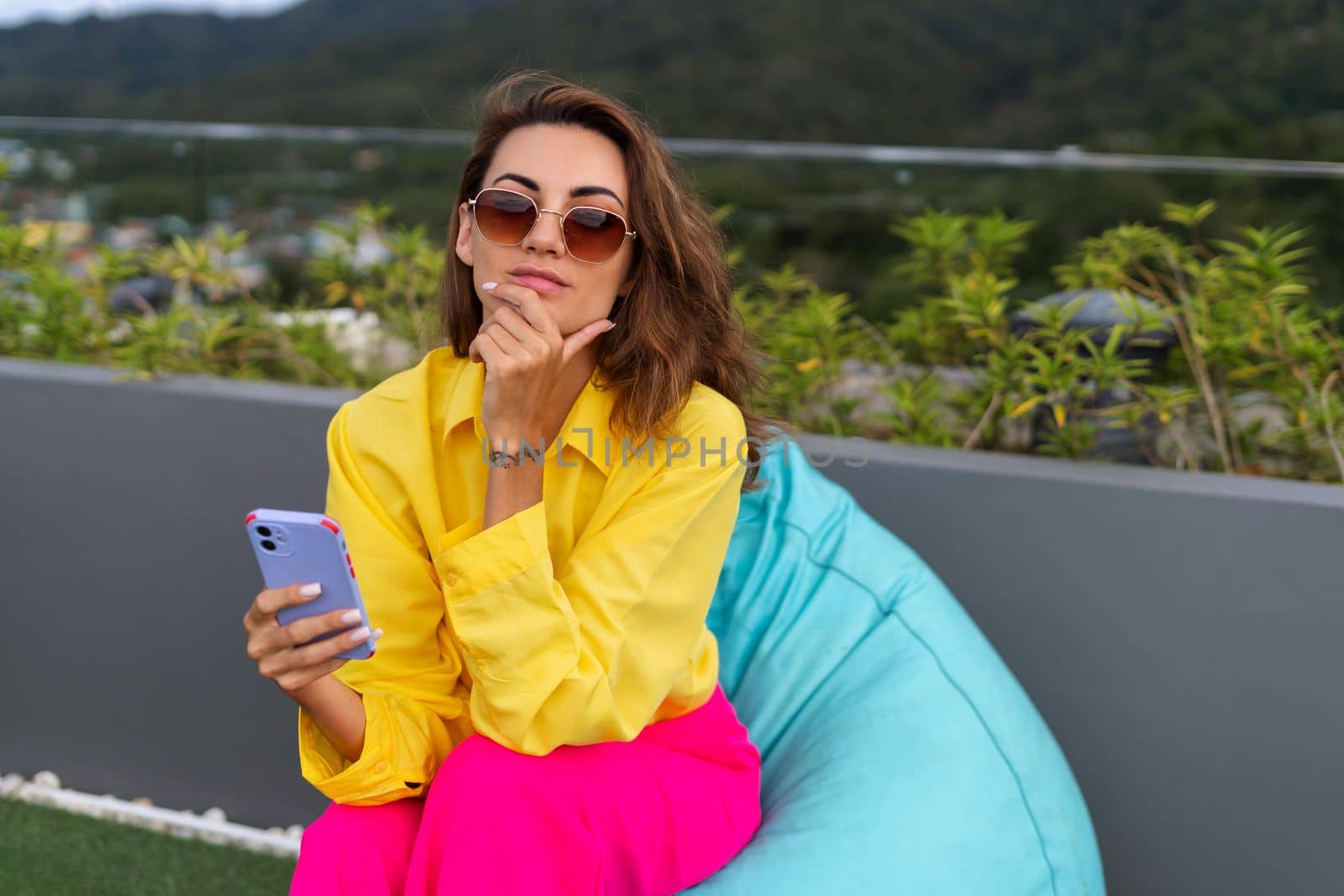 Stylish fit fashion women in bright pink wide leg pants and yellow shirt holding bag trendy sunglasses posing at rooftop terrace tropical view outdoor holding mobile phone