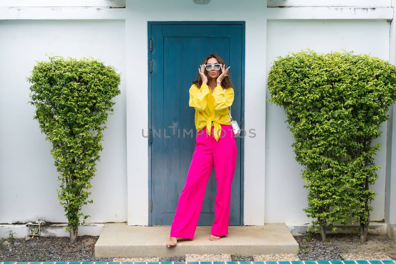 Stylish fit fashion women in bright pink wide leg pants and yellow shirt holding bag trendy mint sunglasses posing at luxury tropical villa by pool outdoor natural day light by kroshka_nastya