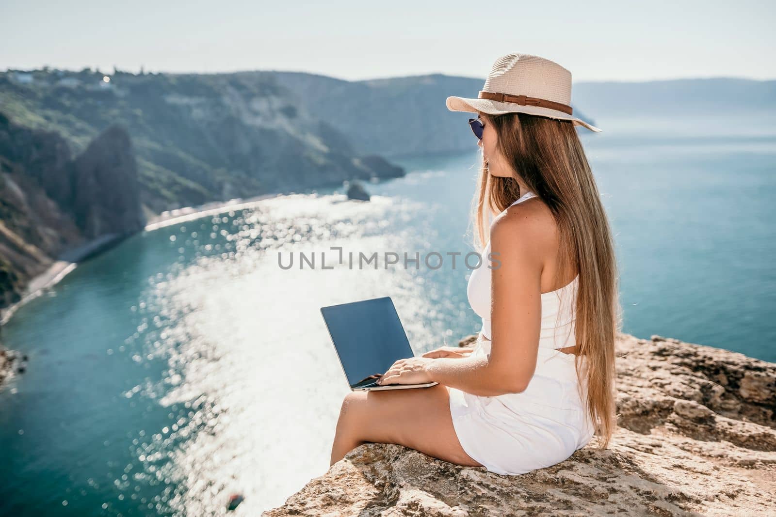 Digital nomad, woman in the hat, a business woman with a laptop sits on the rocks by the sea during sunset, makes a business transaction online from a distance. Freelance, remote work on vacation. by panophotograph