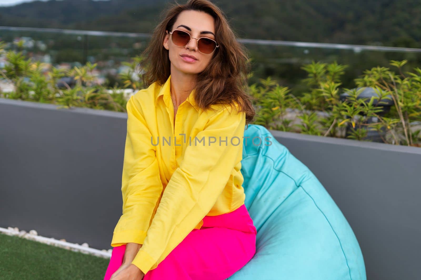 Stylish fit fashion women in bright pink wide leg pants and yellow shirt holding bag trendy sunglasses posing at rooftop terrace tropical view outdoor holding mobile phone by kroshka_nastya