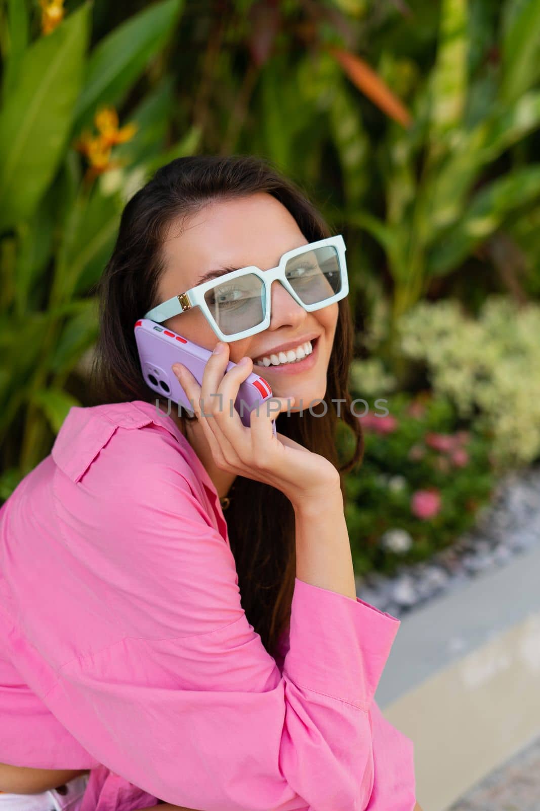 Young beautiful brunette woman in a pink shirt, jewelry on her neck, trendy sunglasses sits on a background of tropical leaves, calls on the phone, smiles by kroshka_nastya