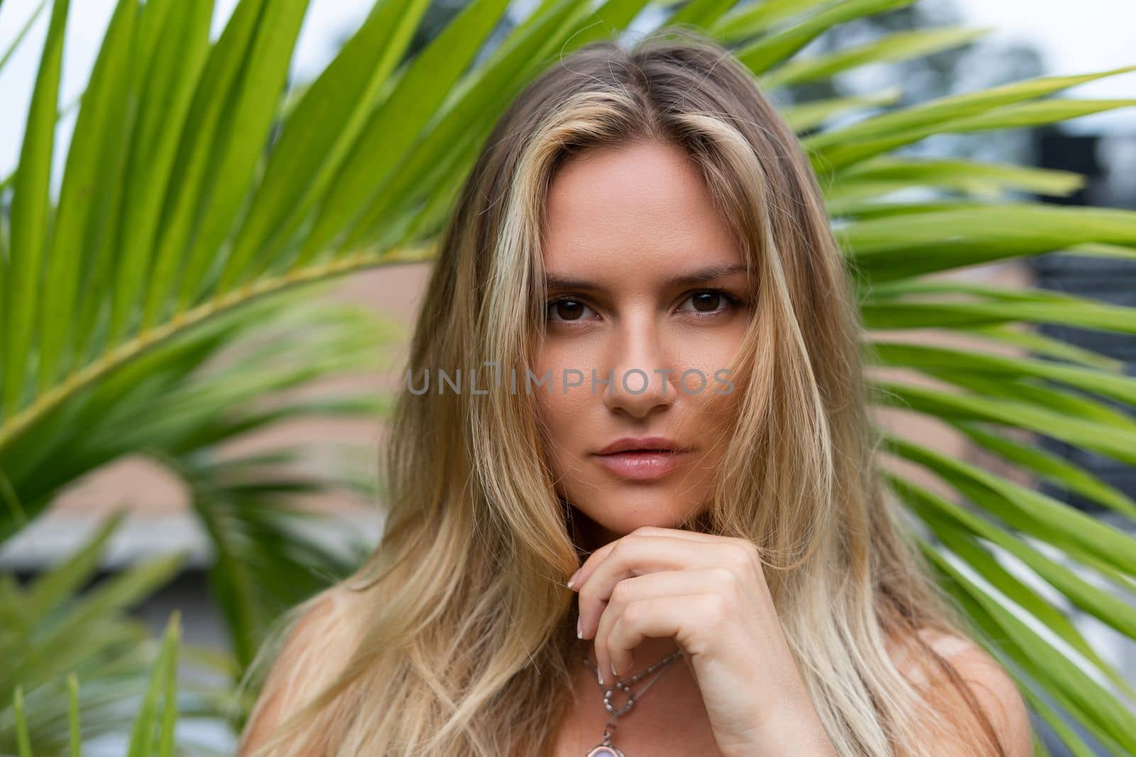 Beautiful young blonde in a top with long hair and smooth clean soft skin, natural beauty, daylight, pendant on neck accessory, posing with palm leaf