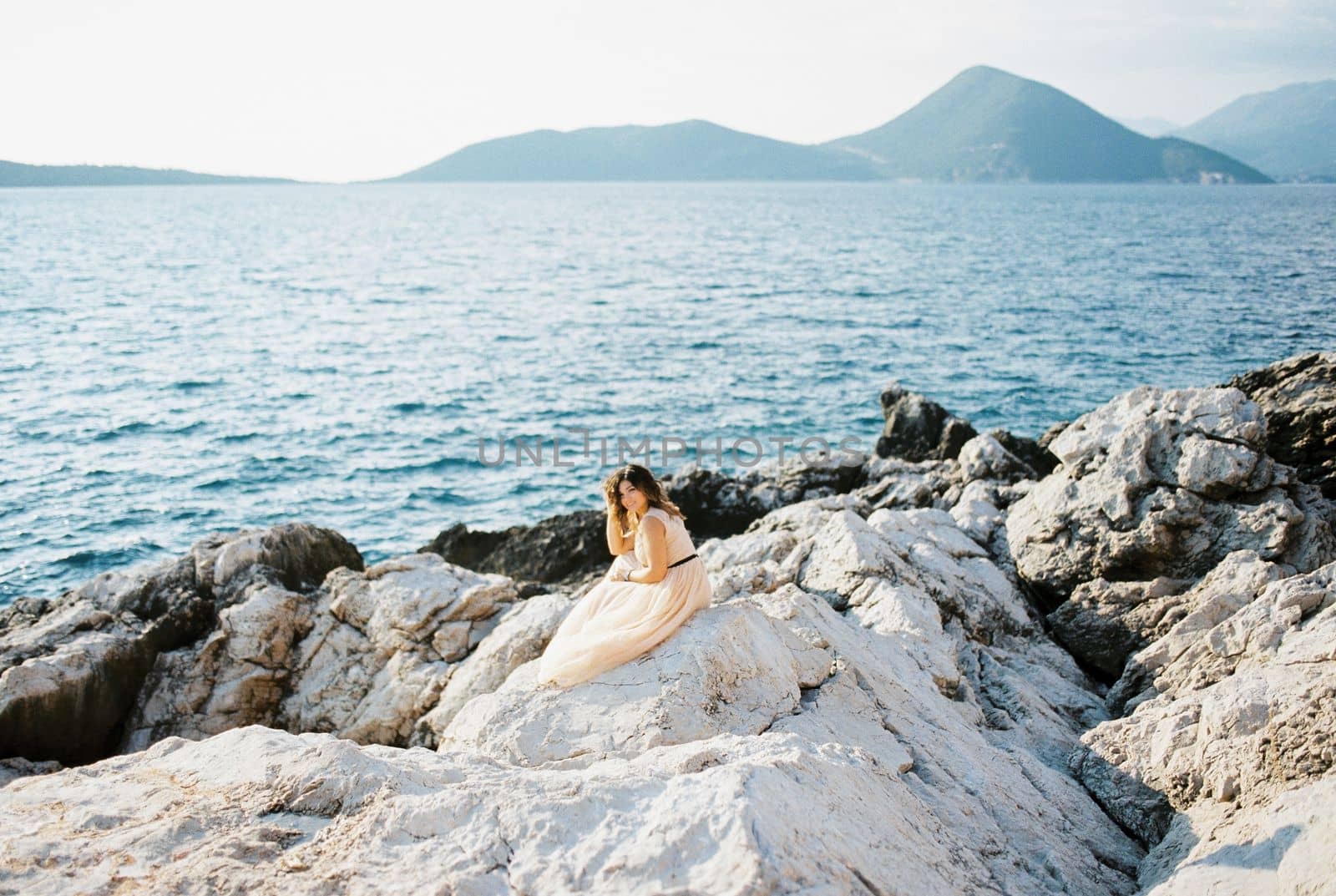 Bride sits on a rocky seashore against the backdrop of mountains, propping her head on her hand. High quality photo