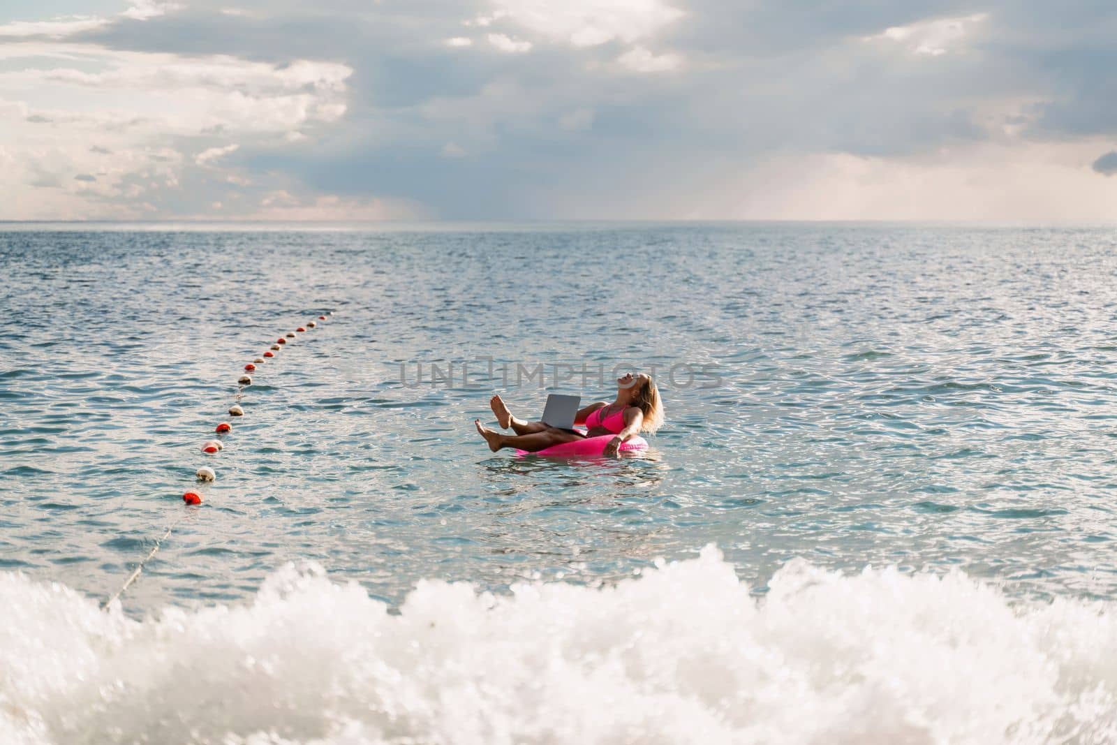 Woman works on laptop in sea. Freelancer, blond woman in sunglases floating on an inflatable big pink donut with a laptop in the sea at sunset. People summer vacation rest lifestyle concept. by Matiunina