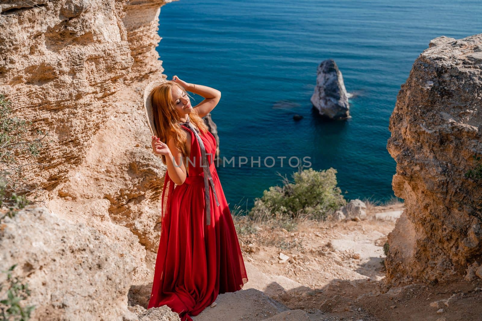 A woman in a flying red dress fluttering in the wind and a straw hat against the backdrop of the sea