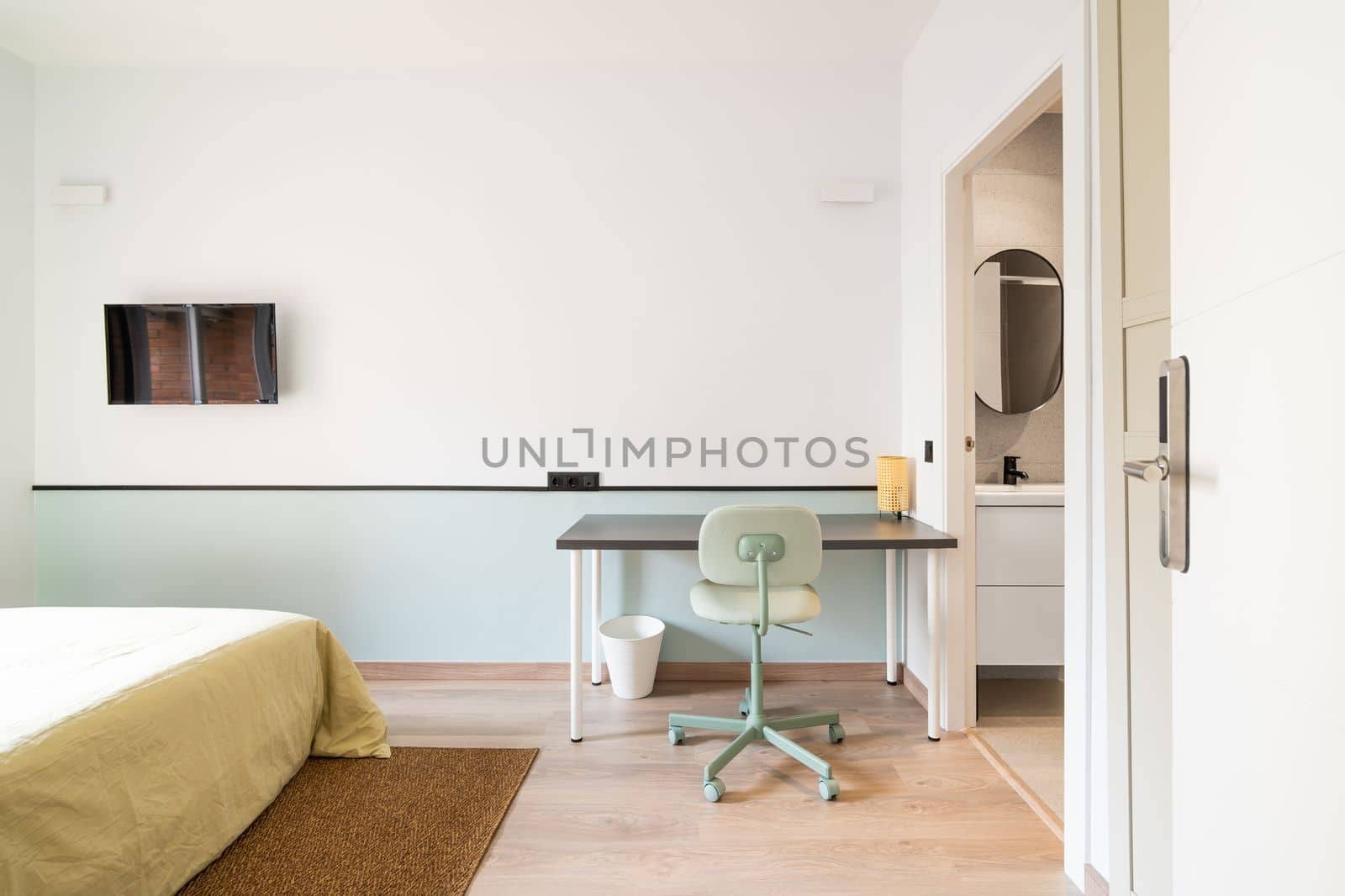 Hotel room with bed for two persons with beige linen. Access to balcony through glass sliding doors. Table against wall with chair for online work. On wall is TV for pleasant evenings watching movie. by apavlin