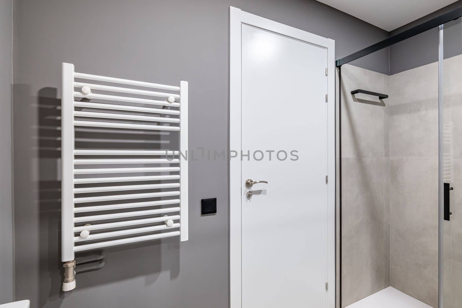 Part of a bathroom with a gray wall. White front door with metal handle. On the wall is a white radiator for drying towels. The shower area is enclosed by sliding glass doors