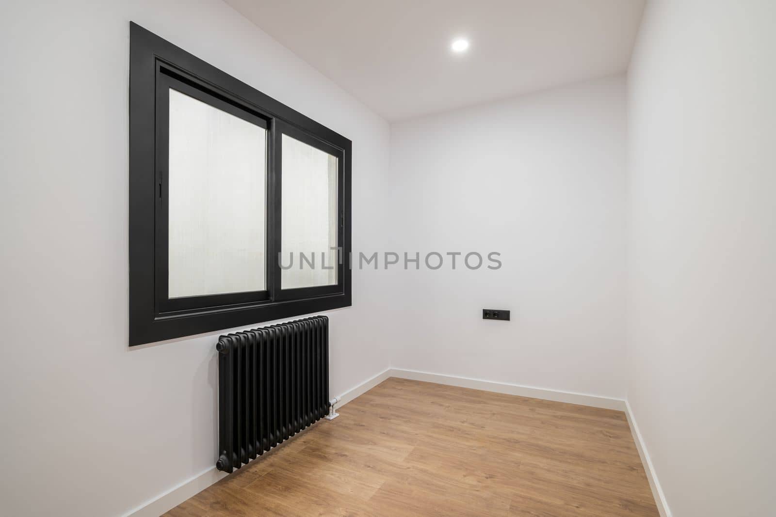 An empty room with a modern version of a plastic window with a black frame and opaque glass. Radiator for heating black. Bright lighting from the ceiling and wooden parquet
