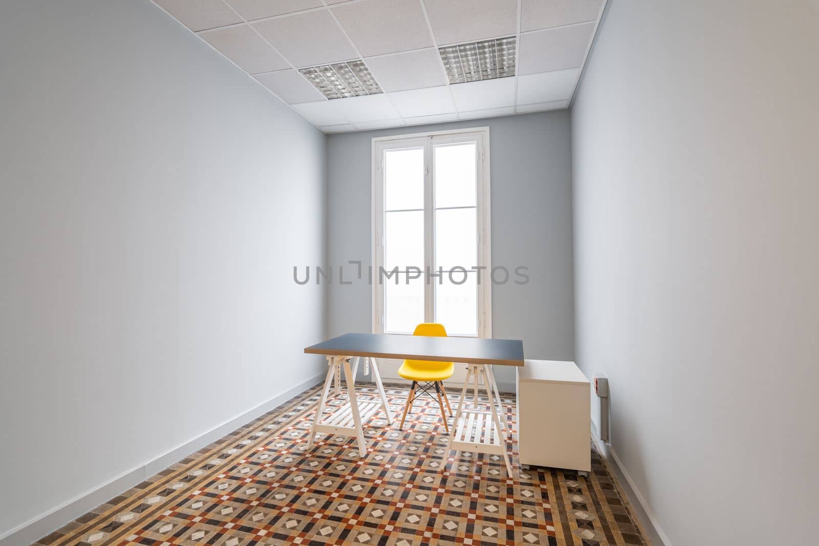 An empty room with gray smooth walls, doors leading to terrace. Floor is made of patterned marble tiles. There is table with gray top a yellow plastic chair with wooden legs in room