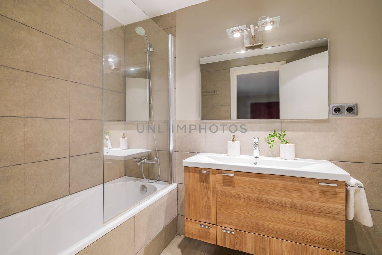 Bathroom in beautiful beige color. Vanity top sink with wooden drawers. Bathtub is fenced with protective glass from splashes. Front door is reflected in square large mirror opposite