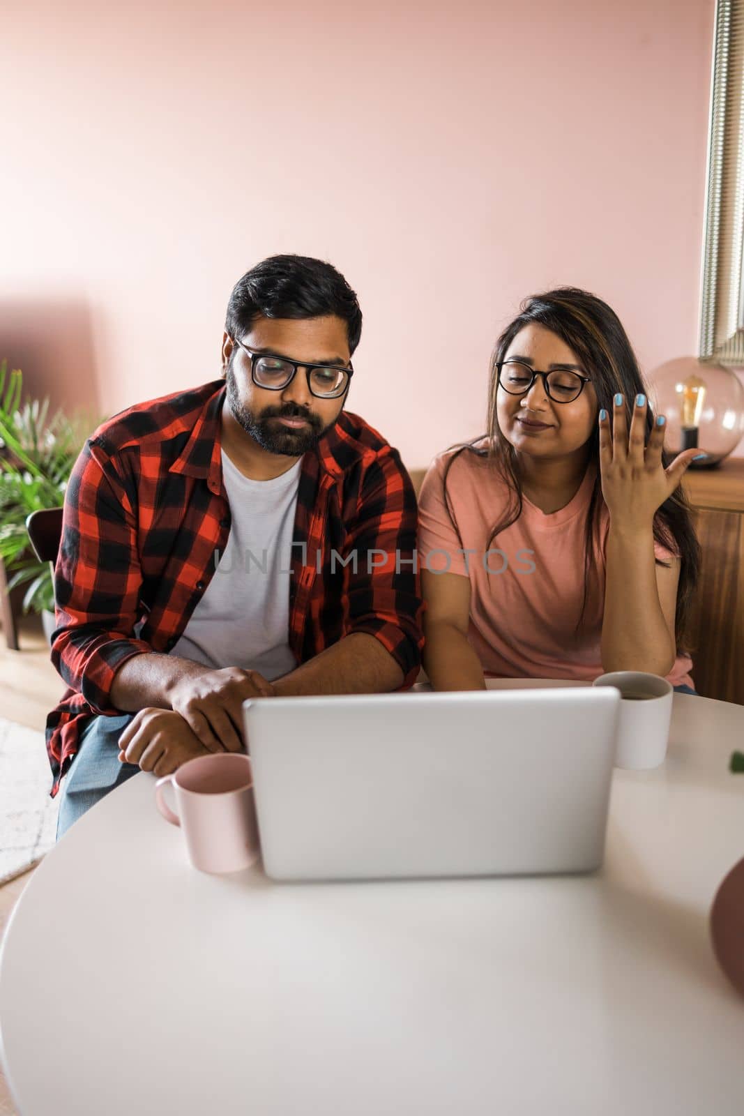 Happy indian family couple cuddle at desk make video call to friends using laptop webcam. Loving young spouses look at computer screen waving hands in good mood greeting parents communicating online via app by Satura86