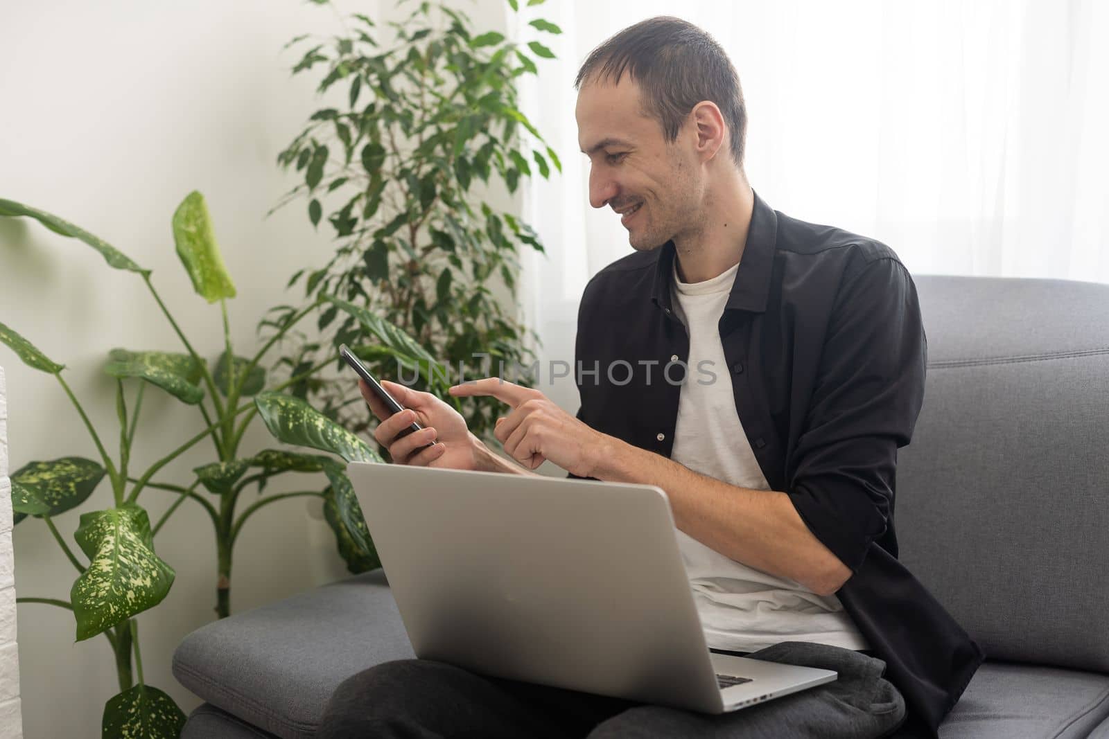 Smiling young man typing on mobile phone while sitting on a sofa at home with laptop computer by Andelov13