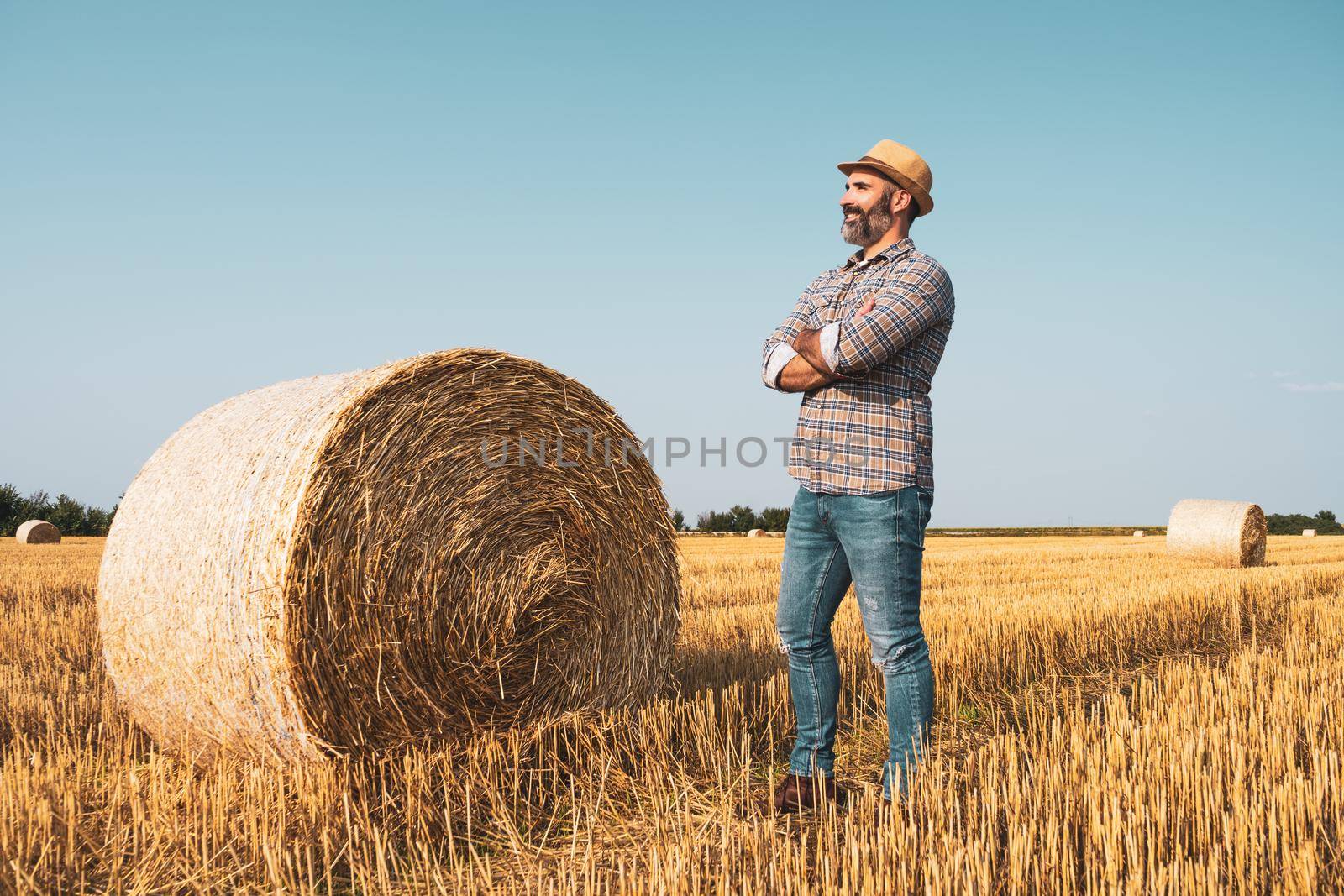 Happy farmer is standing beside bales of hay. He is satisfied because of successful harvesting.