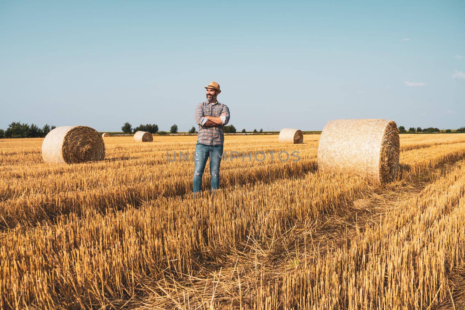 Happy farmer is standing beside bales of hay. He is satisfied because of successful harvesting.