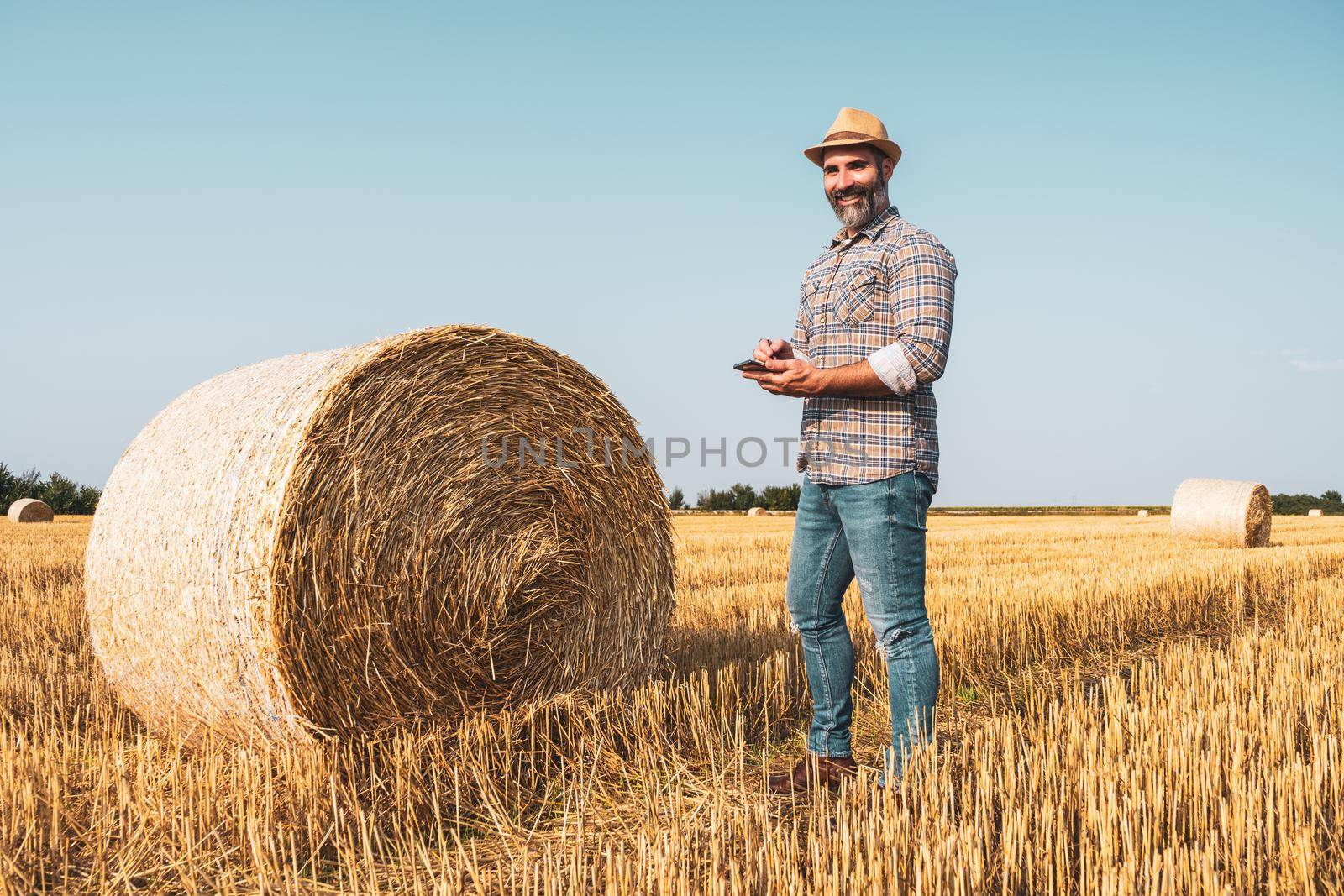 Happy farmer is standing beside bales of hay. He is examining straw after successful harvesting.