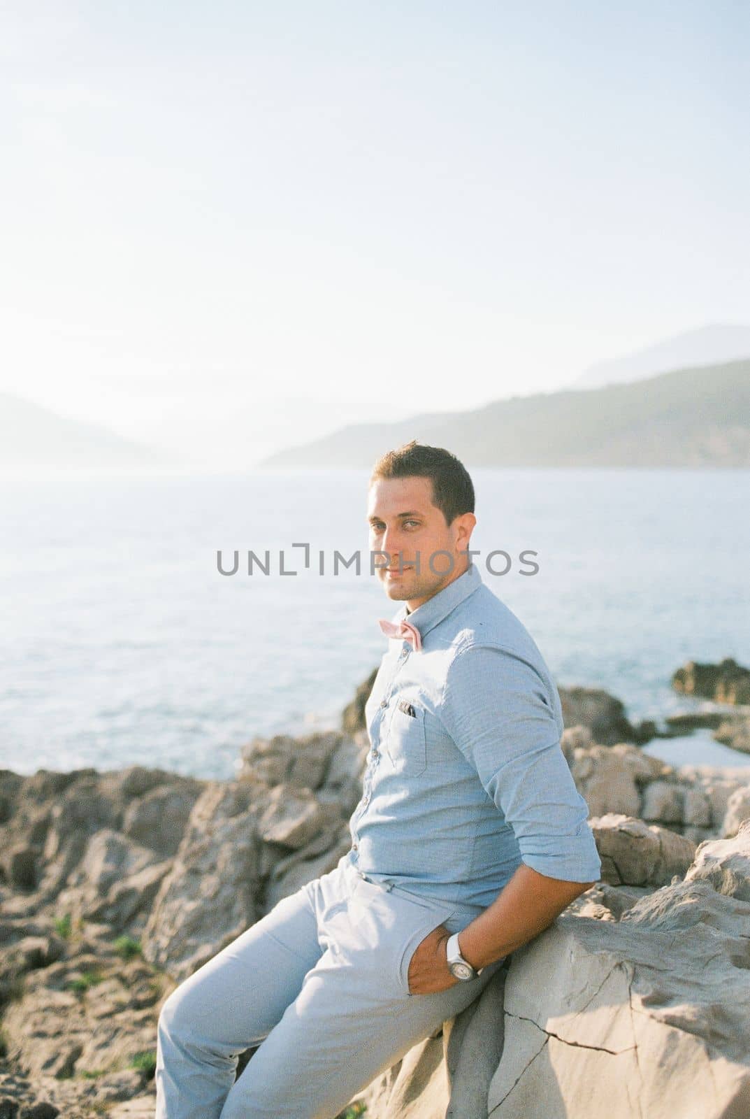 Groom sits on a rocky shore by the sea holding his hands in his pockets. High quality photo