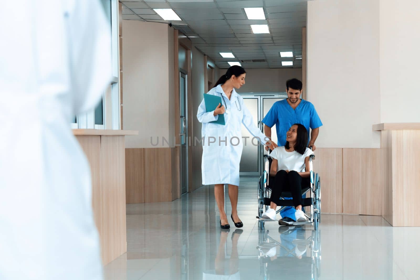 Doctor and male nurse transport a female patient in a wheelchair along sterile hospital corridor. Health care and nursing care for disabled handicapped patient in the hospital concept.