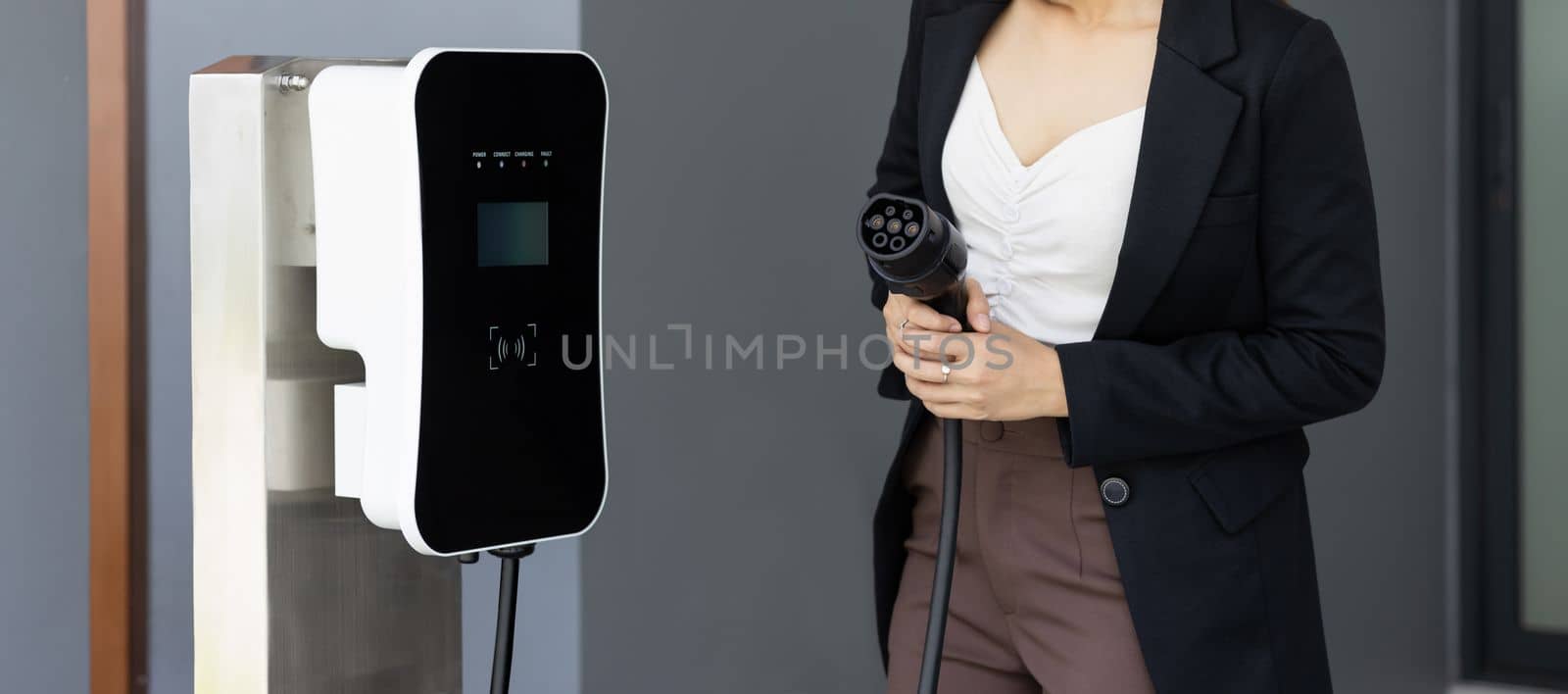 Closeup progressive asian woman holding EV plug at home charging station. by biancoblue